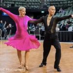 Ceasar Chong dancing with a student. Caesar is black pants and shirt, his student is in bright pink. They are dancing a high-energy Latin dance.